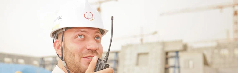 Health and safety inspector wearing a hard hat and using a walkie-talkie on a construction site.