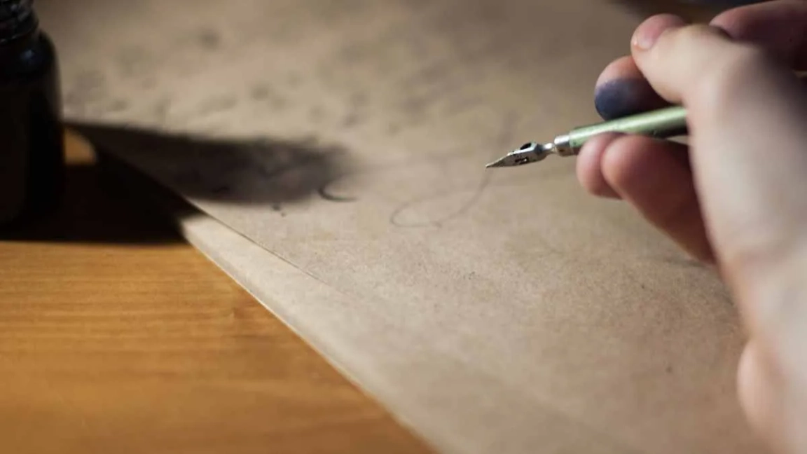 A hand holding a fountain pen over a piece of paper filled with calligraphic writing and an ink pot to the side
