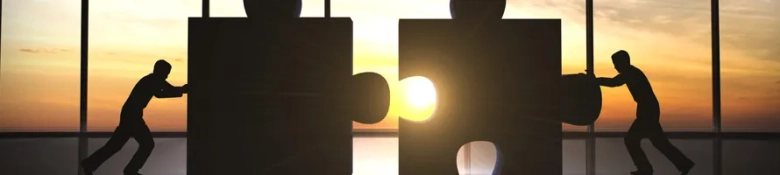 Silhouettes of two people pushing large puzzle pieces together against a sunset background, symbolizing collaboration in marketing management.