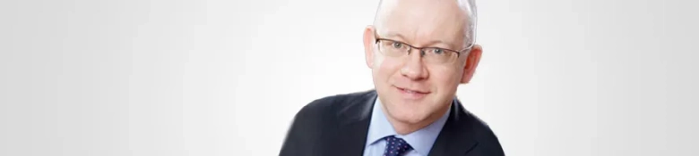 Professional headshot of Shaen Catherwood, wearing glasses and a suit with a blue tie.