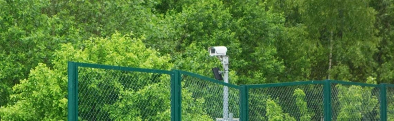 A security camera atop a post. There are trees in the background and a green fence in the foreground.