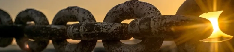 A chain with the sun shinning through one of the holes of one of the chain links.