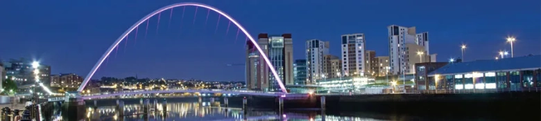 Nighttime view of the illuminated Gateshead Millennium Bridge and modern buildings along the riverbank.