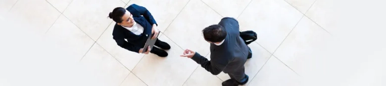 Two professionals in business attire engaged in a discussion, one holding a tablet, viewed from above.