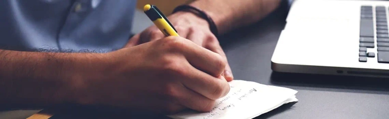 Close-up of a person's hand writing notes on a pad of paper next to a laptop on a desk.