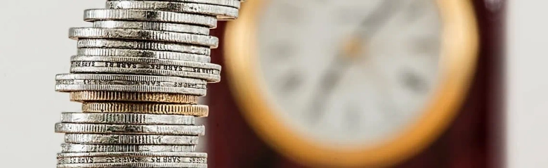 Stack of coins in focus with a blurred clock in the background, symbolizing time and money management.