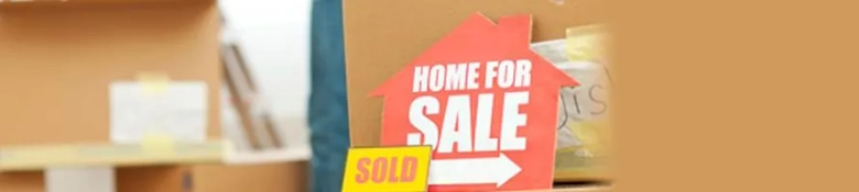 Red 'Home for Sale' sign with 'Sold' sticker and blurred moving boxes in the background.