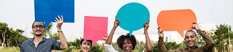 Four diverse people smiling and holding up colorful speech bubble-shaped signs outdoors.