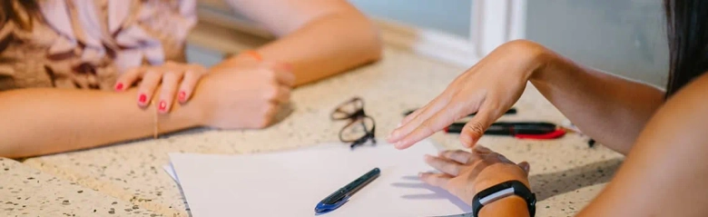 Close-up of a counselling session with hands in focus, one person gesturing while the other listens, with paper and pen on the table.