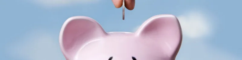 Hand inserting a coin into a pink piggy bank against a blue sky background, symbolizing savings and financial management.