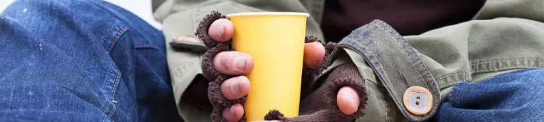 Close-up of a person in worn gloves holding a yellow cup for donations, symbolizing homelessness and the need for support.