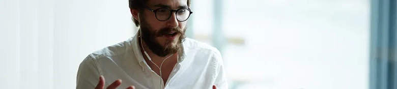 A bearded man wearing glasses and a white shirt using headphones on a video call.