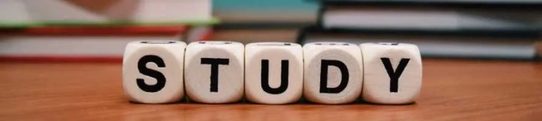 Individual dice spelling out "study" in capitals, sitting on a table with red and blue books behind it