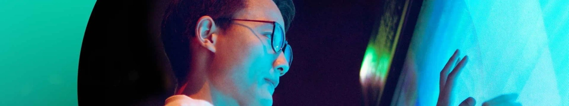 Profile of a person with glasses looking at a brightly lit screen, reflecting blue and green light on their face.