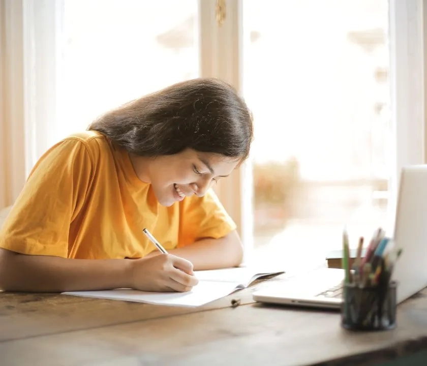 A young woman in an orange t-shirt writing in a notepad. She has a laptop open in front of her.