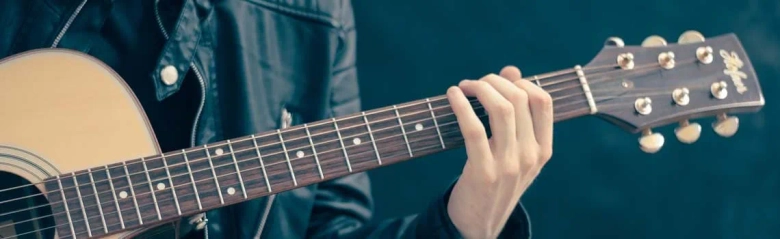 A musician in a leather jacket playing a wooden acoustic guitar: private music teachers and tutors teach music lessons in various instruments