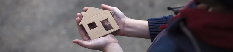 Hands holding a cardboard cutout of a house, symbolizing homelessness and the need for shelter.