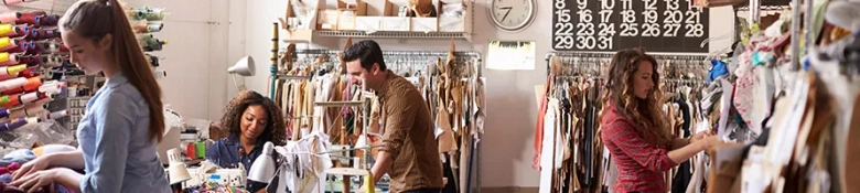 Three people engaged in various activities in a fashion design studio, with fabrics and clothing samples around them.