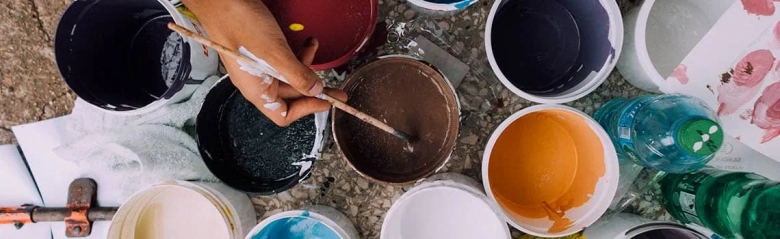 A painter's hand dipping a brush into a tub of brown paint.