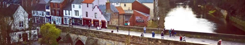 Panoramic view of Durham with people walking along the riverbank, historic buildings in the background, and a water feature spraying into the air.