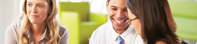 Three professionals in a meeting, with a focus on a smiling man in a tie engaging in a discussion with colleagues.