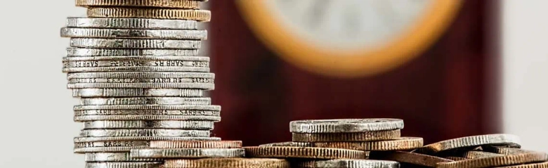 Stacked coins in various denominations with a blurred clock in the background.