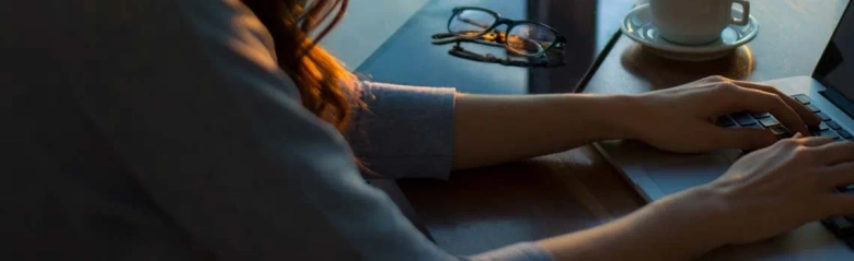 Close-up of a person's hands typing on a laptop with eyeglasses and a coffee cup in the background.