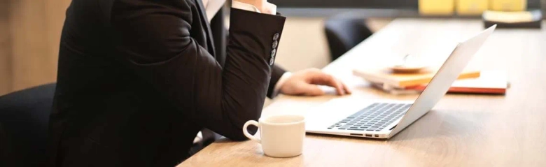 Professional working at a desk with a laptop and a notepad, with a coffee cup on the side.