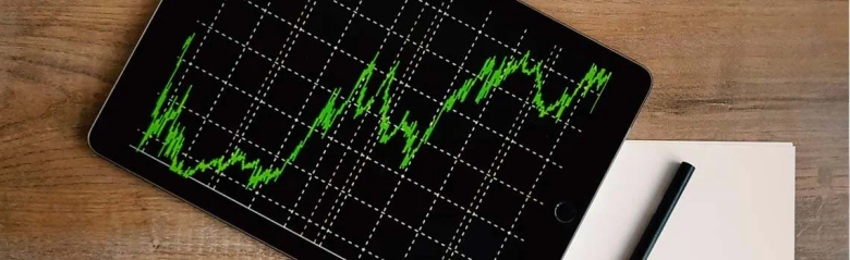 Tablet displaying stock market graph next to a notepad and pen on a wooden surface.