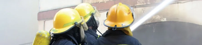 Firefighters in yellow helmets using a hose to extinguish a fire.
