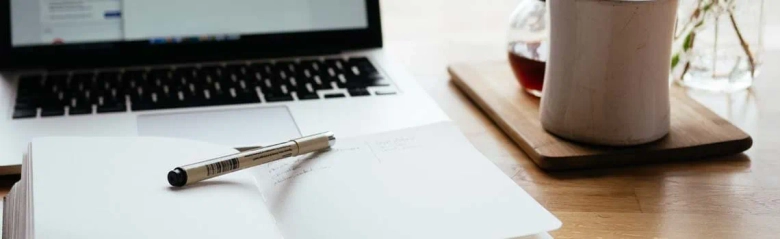 Work desk with an open notebook, pen, laptop, and a cup of coffee.