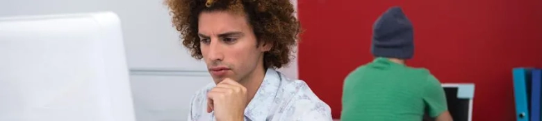 Man with curly hair pondering in front of a computer screen with another person working in the background.