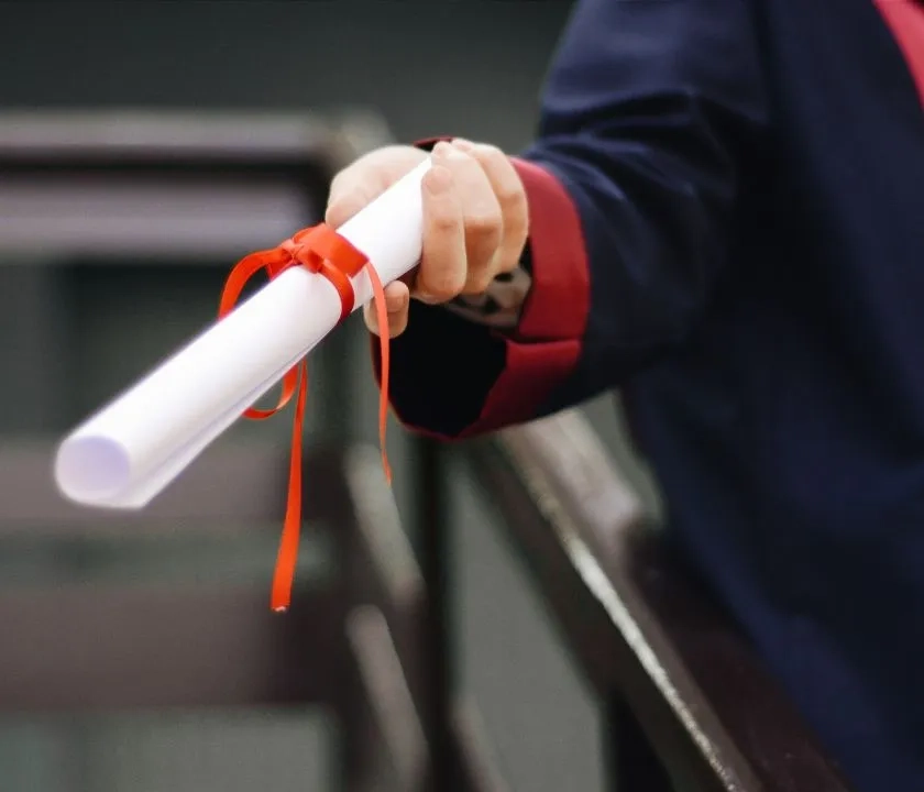 A graduate in a black gown holding out a rolled-up degree certificate tied with a red ribbon. 