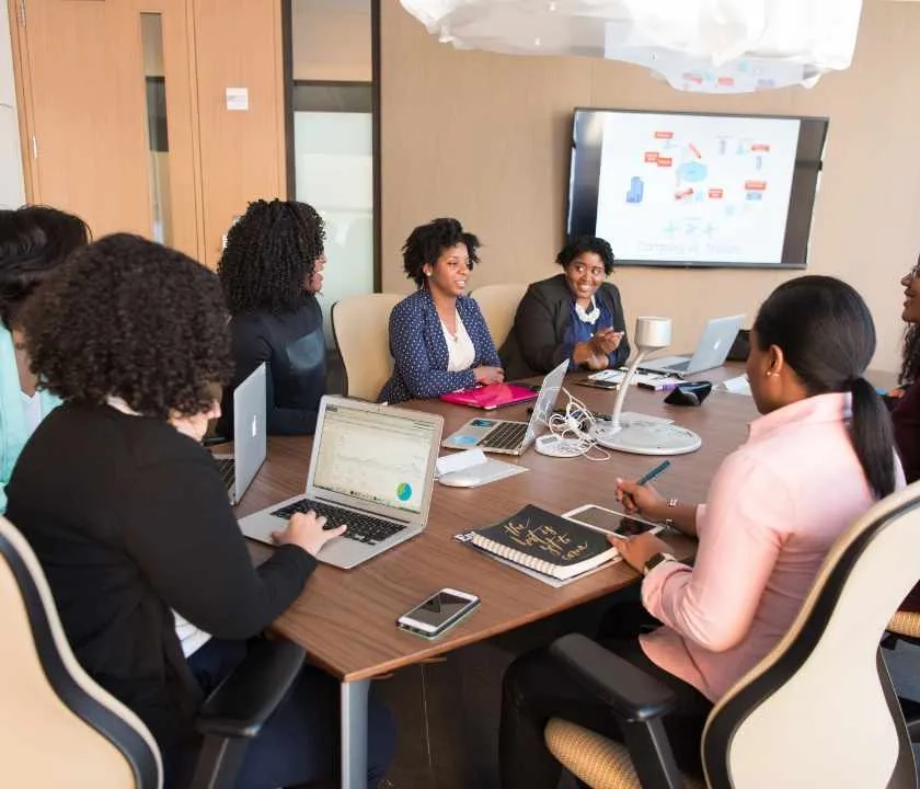 A collection of professionals sitting around a meeting room table with a presentation on screen 