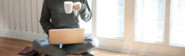 A writer sitting cross-legged on the floor with a drink in a mug, writing on a laptop