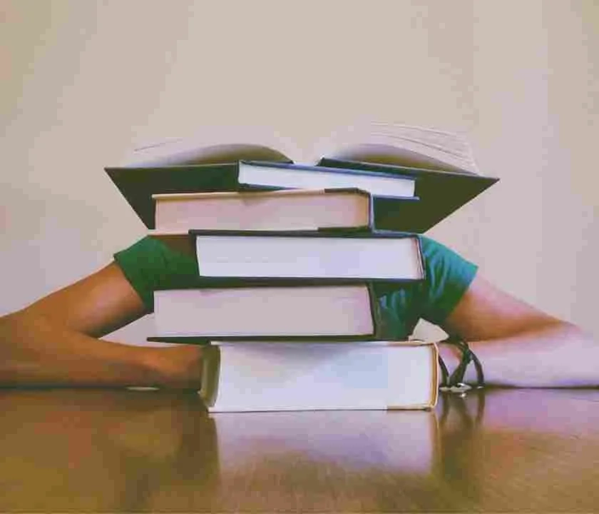 A pile of books with a student sitting behind them. The books drawf the person, so that only their arms are showing