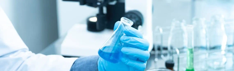 A chemical engineer's hand wearing a blue glove and holding a conical flask containing blue liquid.