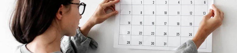 Woman in glasses pinning a blank monthly calendar on a wall.