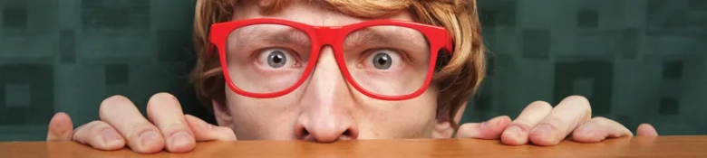 Person with red glasses peeking over the top of a wooden barrier with a surprised expression.