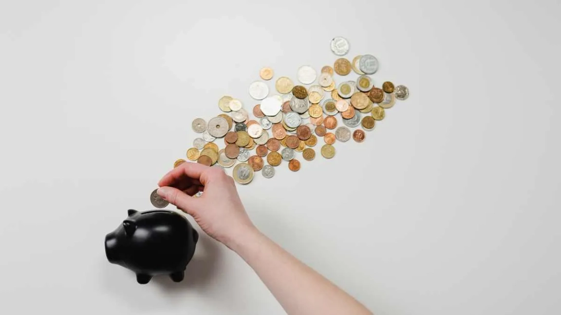 A hand putting money into a piggybank, with a scattering of coins nearby 