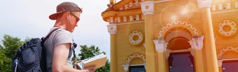 Tour guide wearing a cap and backpack reading a guidebook in front of a historic building.