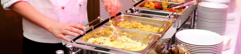 Chef serving food at a buffet with various dishes and stacked plates.