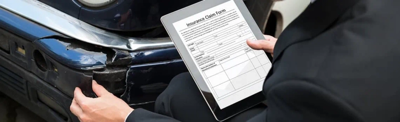 Insurance claims inspector examining damaged car and holding a digital tablet displaying an insurance claim form.