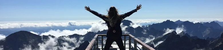 A woman standing on a mountain viewpoint looking onto other mountains with her arms outstretched in jubilation.