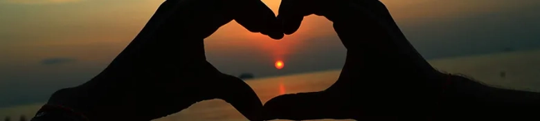 Hands creating a heart in front of a sunset over tropical beach