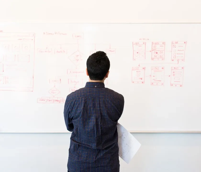 A man facing a white board. There are calculations written on the whiteboard. The man has an inquisitive gesture. 